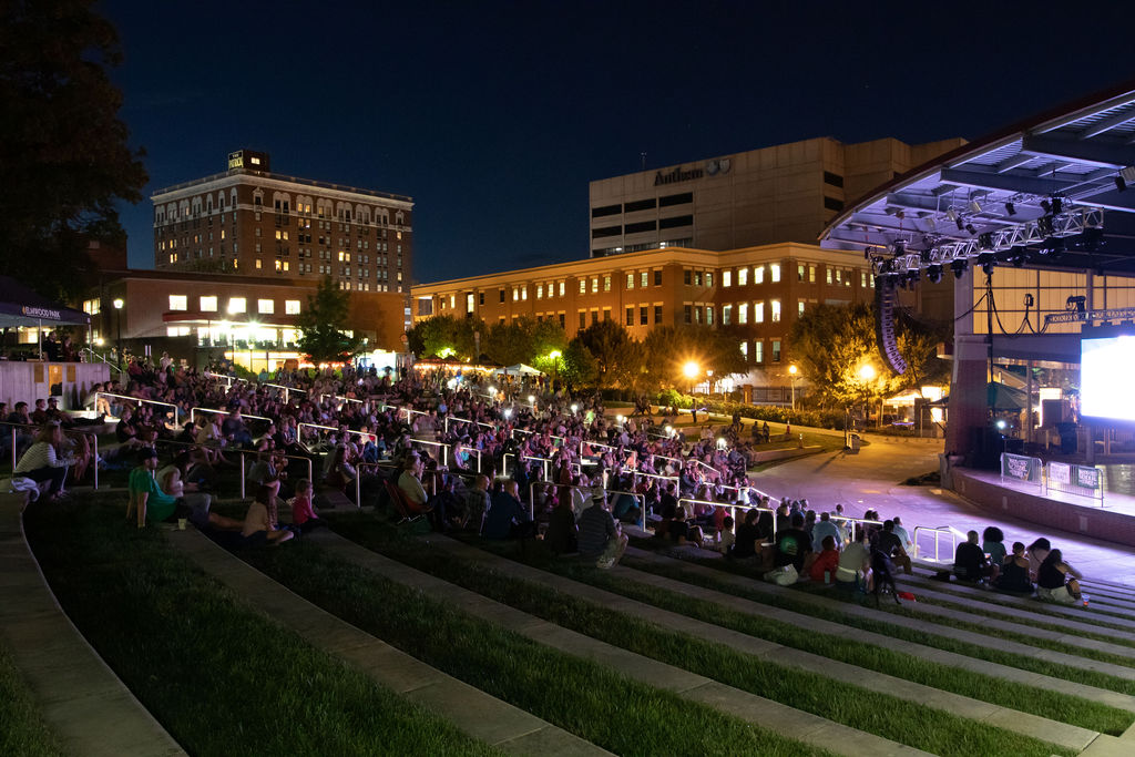 Outdoor amphitheater at night showing a movie with several people on the terraced seating facing the amphitheater.